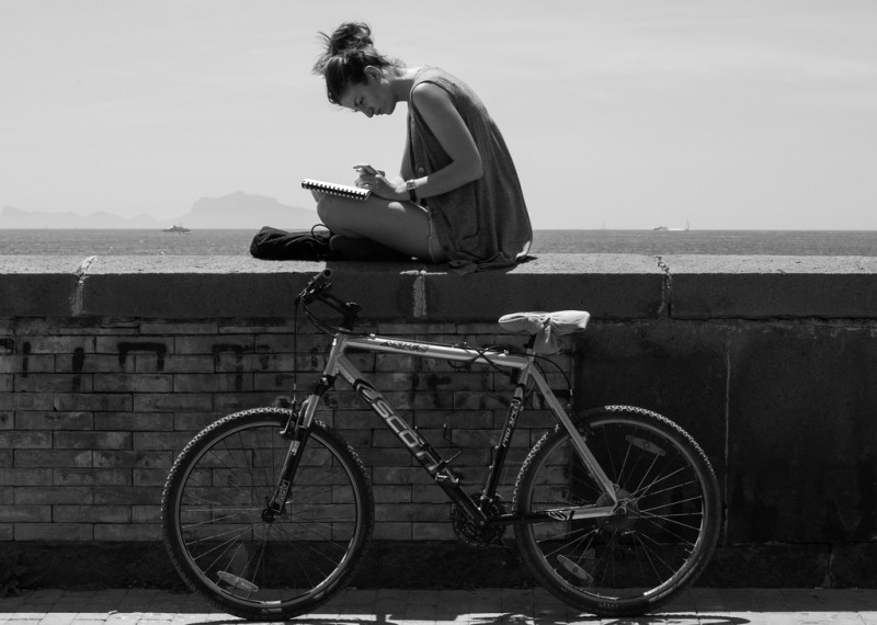 Una mujer escribe a mano en un cuaderno y debajo, apoyada en un muro, hay una bicicleta