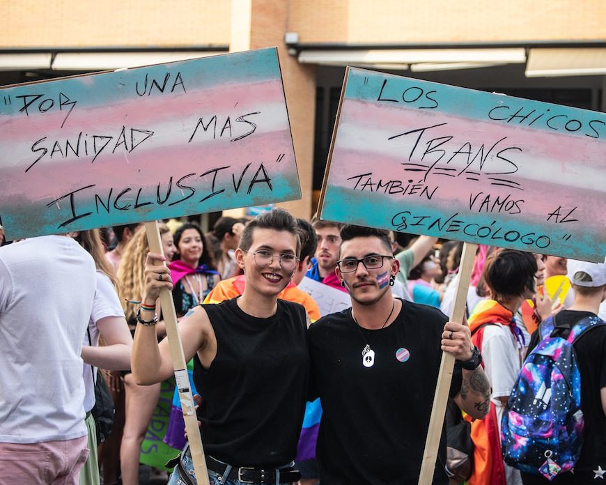 Dos personas con carteles con los lemas "Por una sanidad más inclusiva" y "los chicos trans también vamos al ginecólogo"