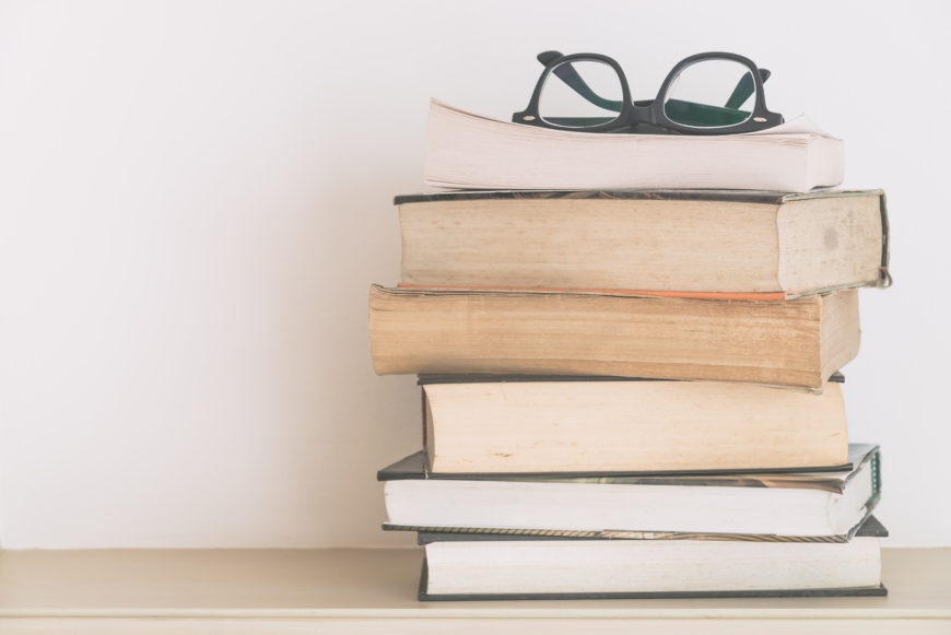 En la imagen se ven seis libros apilados sobre una mesa y unas gafas de ver cerradas encima de ellos.