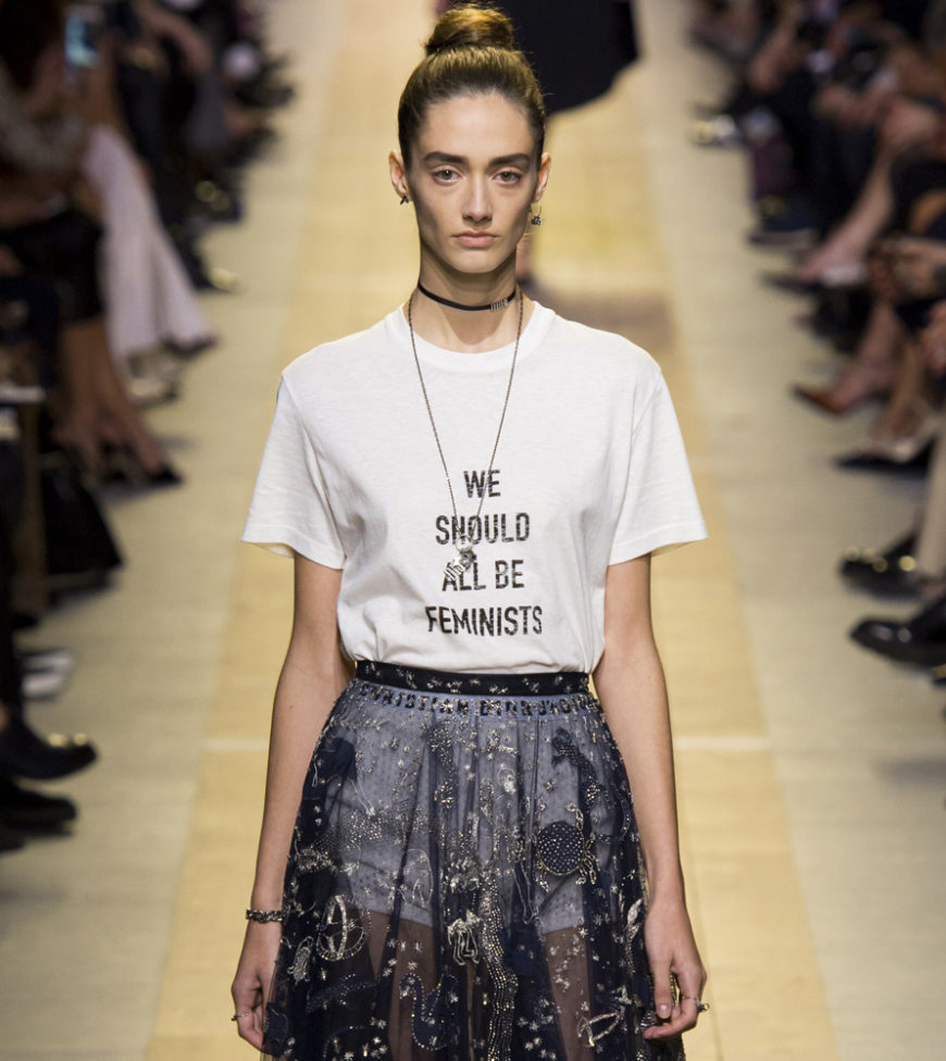 Foto de una modelo en una pasarela, luciendo una camiseta blanca en la que pone "Todos deberíamos ser feministas" en inglés.