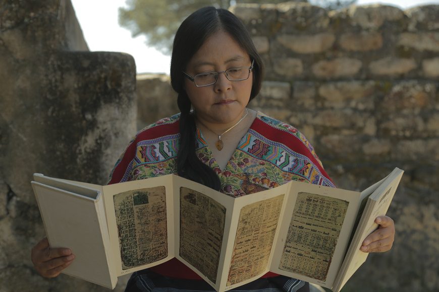 Un a mujer lee un trípico. Lleva el pelo recogido en con una trenza echada para un lado y gafas.