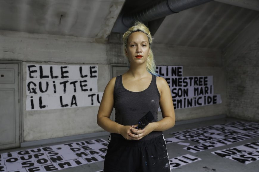 en mitad de la imagen una mujer posa, con un pincel en la mano, al fondo se ven carteles que ha hecho. Francia