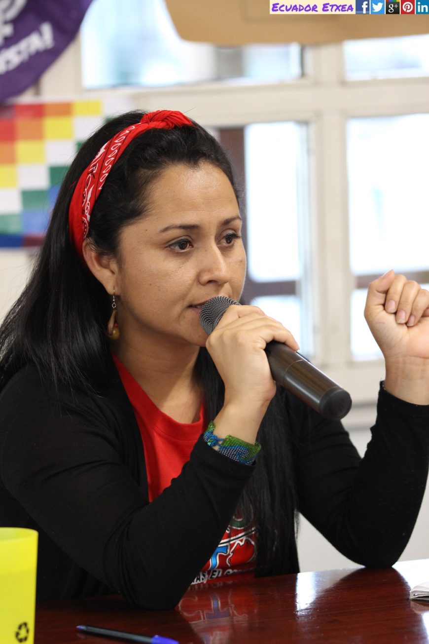 una mujer sentada habla con un micro en la mano. Se ve solo medio cuerpo. tiene el pelo suelto y moreno y un cintillo rojo