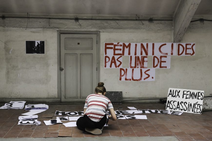 una sala casi vacía, con un cartel con letras rojas y de rodillas y de espaldas una mujer hacer un cartel en el suelo. Francia