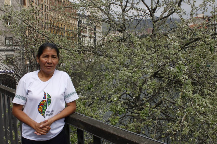 una mujer con camiseta blanca, posa, de medio cuerpo, apoyada en una barandilla. Al fondo un árbol que tapa edificios de Bilbao