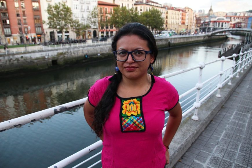 en el centro de la imagen, y de medio cuerpo, una mujer, con camiseta de manga corta, las manos en los bolsillos traseros del pantalón, gafas y el pelo recogido. Al fondo, la ría de Bilbao