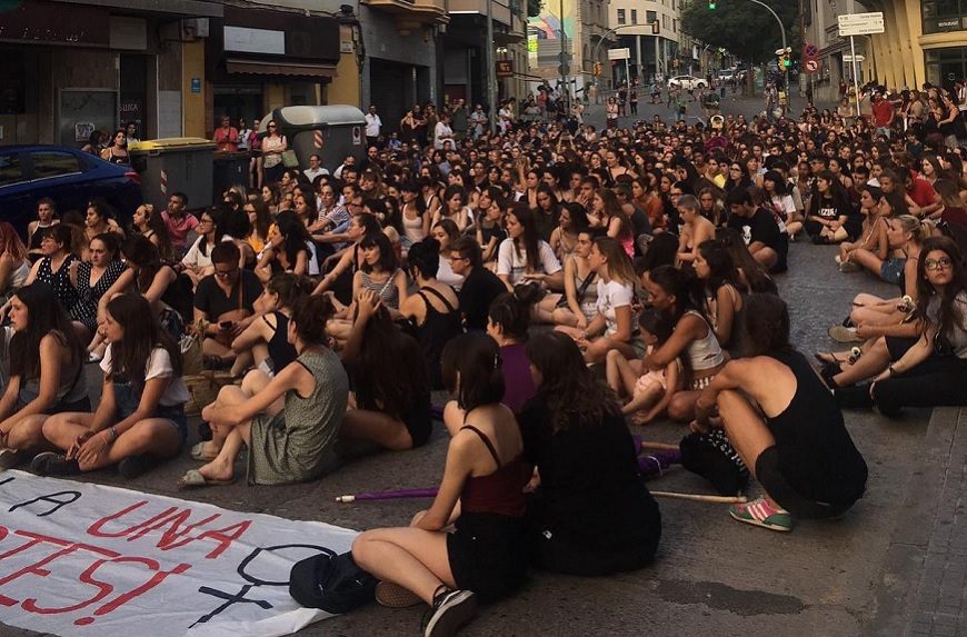 Un grupo de mujeres está sentado en el suelo en medio de la calle de una ciudad con una pancarta