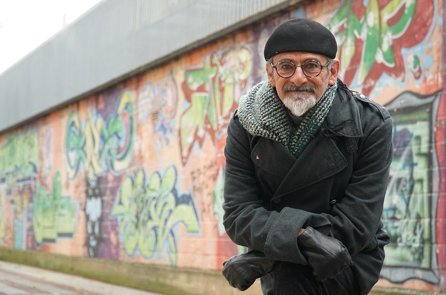 un hombre posa para la foto, frente a una pared pintada. Va con gorrito, vestido de negro, gafas redondas y barbita blanca