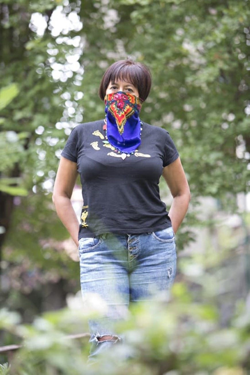 una mujer posa, en medios de vegetación, con un pañuelo de colores que le tapa media cara, de nariz hacia abajo