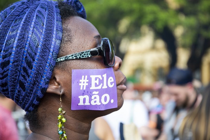 una mujer negra de perfil, con pañuelo en la cabeza, gafas de sol y un cartel en la cara que pone Ele Nao