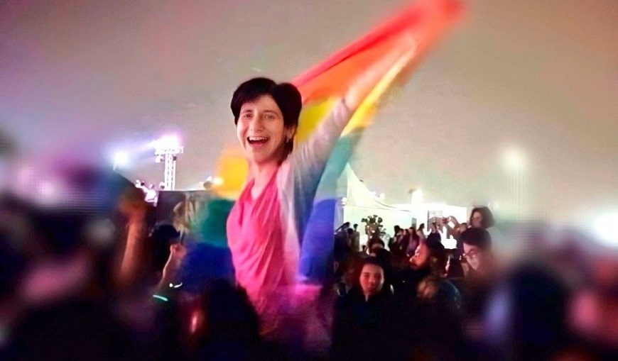 una chica entre una multitud se levanta sonriente con la bandera LGTBI