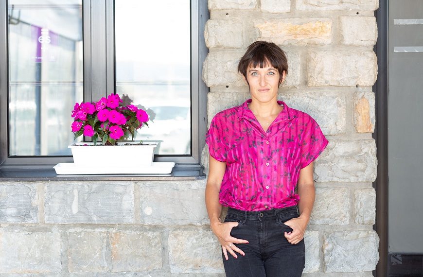 una mujer posa en una fachada de una casa en la que hay una ventana con una maceta de flores. el color de su camisa es el mismo que el de las flores, rosa fucsia