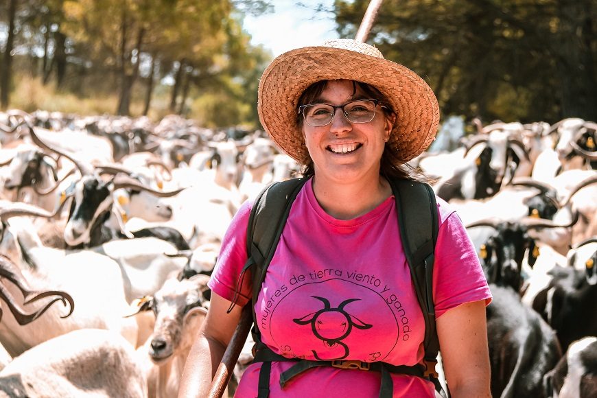 en primer plano una ganadera de medico cuerpo y con sombrero de paja y al fondo se ven sus cabras