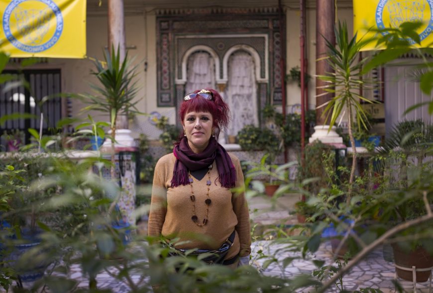 Una mujer posa entre plantas. Tiene el pelo teñido de rojo, lleva unas gafas en el pelo, un pañuelo en el cuello y collares