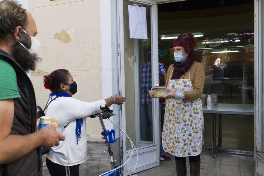 Una mujer, a la puerta de un establecimiento, con mascarilla y un mándil, tiene un táper en la mano y lo lleva a una fila de gente