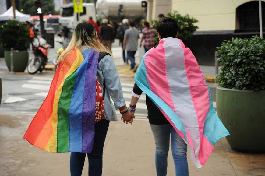 dos personas caminando cogidas de la mano con la banderas a la espalda lgtb y trans