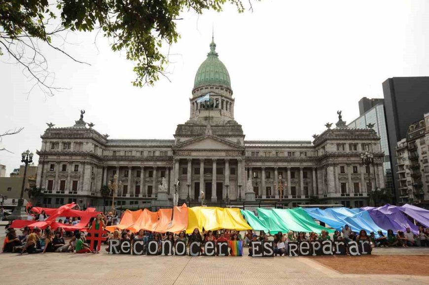 un montón de gente delatne de un edificio público forman la bandera arcoiris y las palabras reconocer es reparar