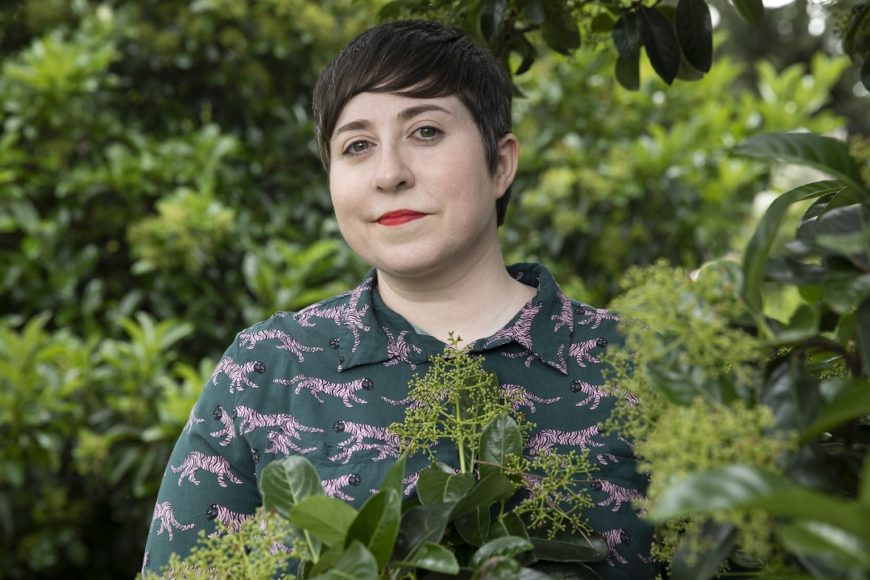 una mujer posa, de medio cuerpo, entre plantas y hojas verdes