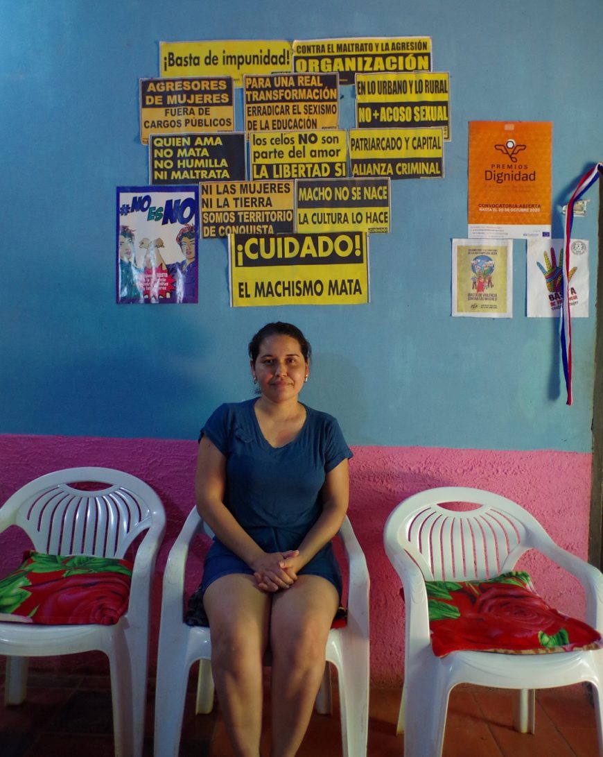 una mujer sentada en una silla de terraza de bar, dentro de un local con las paredes pintadas azul y rosa y con carteles con frases feministas pegadas