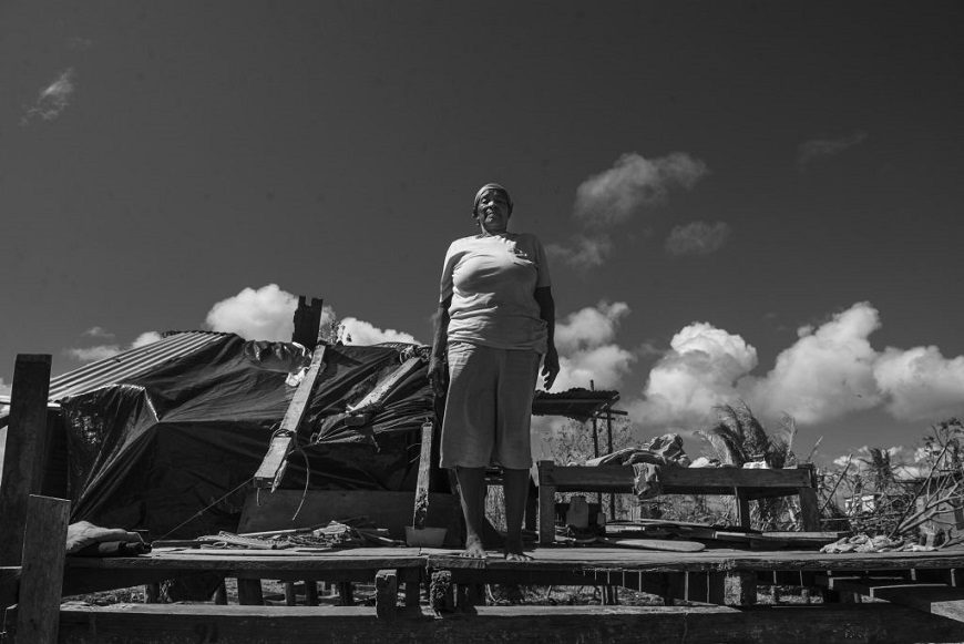 foto contrapicada de una mujer que posa delante de los restos de madera de una casa que está reconstruyendo. foto en blanco y negro