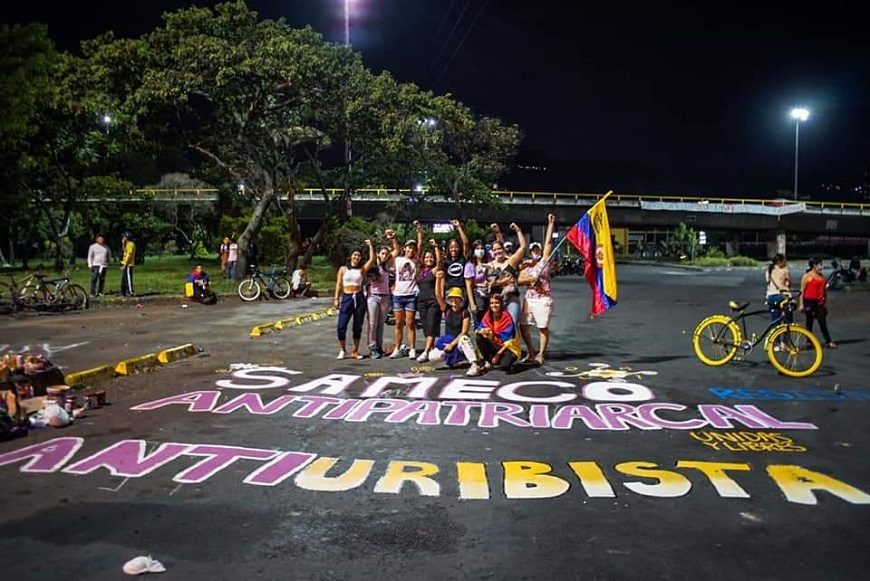 un grupo de mujeres con brazos en alto y la bandera de Colombia posan ante pintadas en el suelo contra Uribe