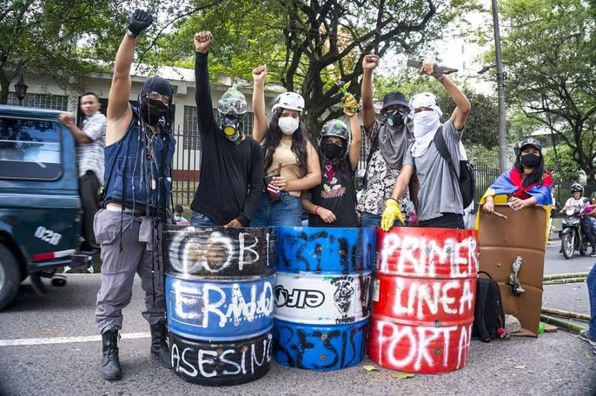 varias personas con la cara tapada y los brazos en alto detrás de unos bidones