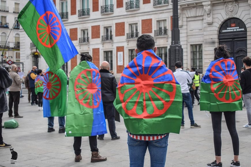 un grupo de gente, en una concentración, separadas por la covid, en la puerta del sol de madrid. Están de espaldas y llevan la bandera gitana a modo de capa