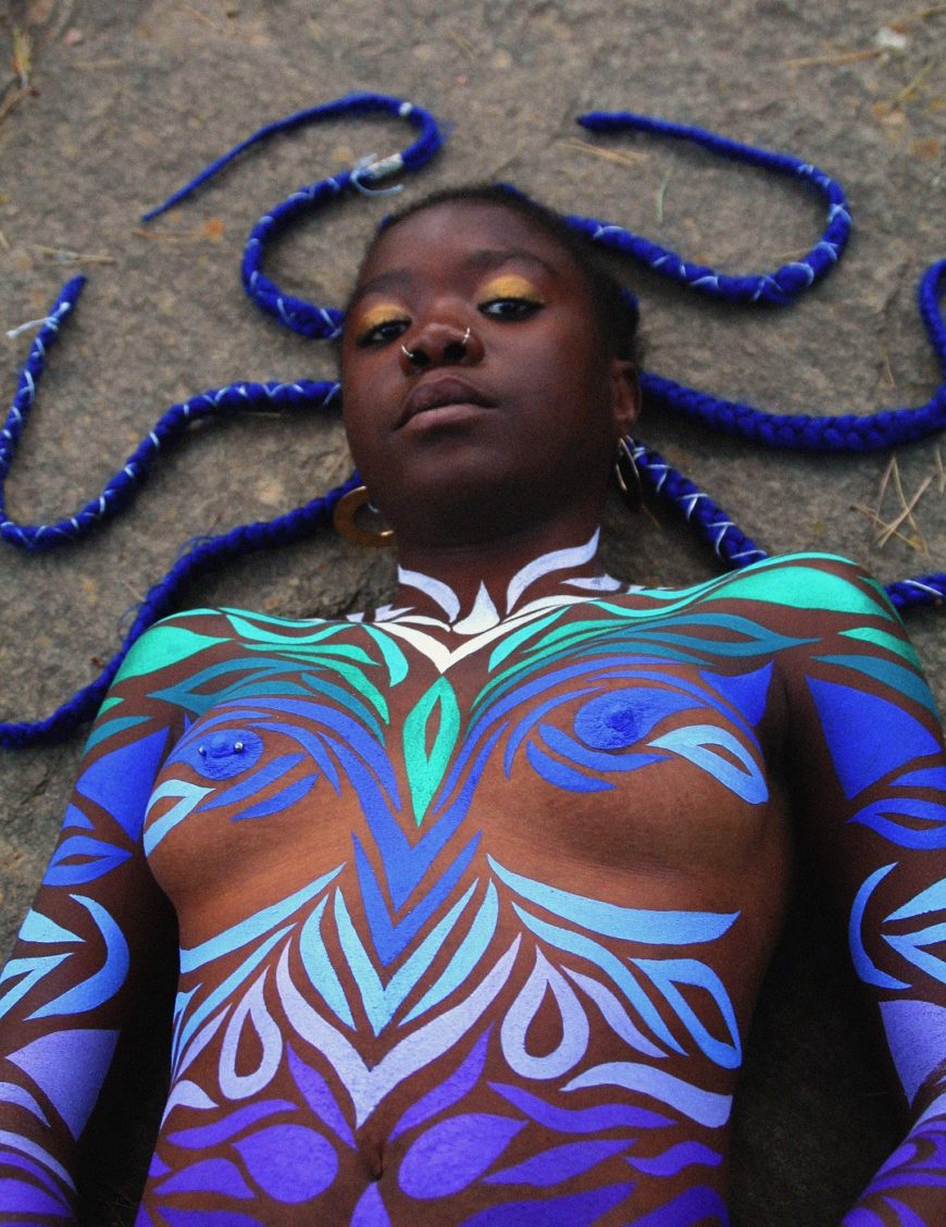 una mujer negra, desnuda, tumbada de medio cuerpo, que está pintado y con unas rastas azules extendidas por el suelo