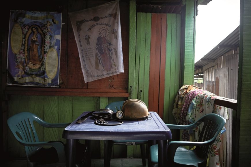 una mesa y unas sillas de plástico, dentro de una casa de madera con la ventana abierta. Hay una imagen de la virgen colgada