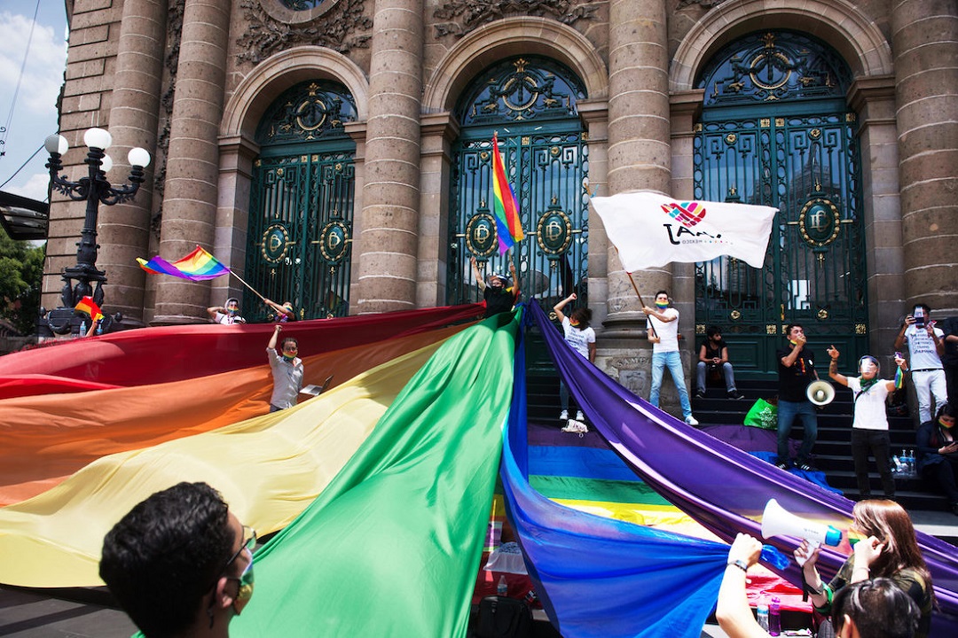 una protesta frente a un edificio importante de México donde se ve una gran bandera arcoiris