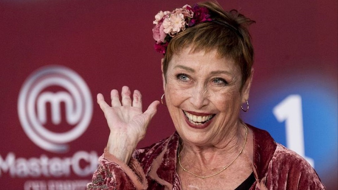 una mujer saluda sonriente con la mano. Lleva pelo corto y una flor en el pelo